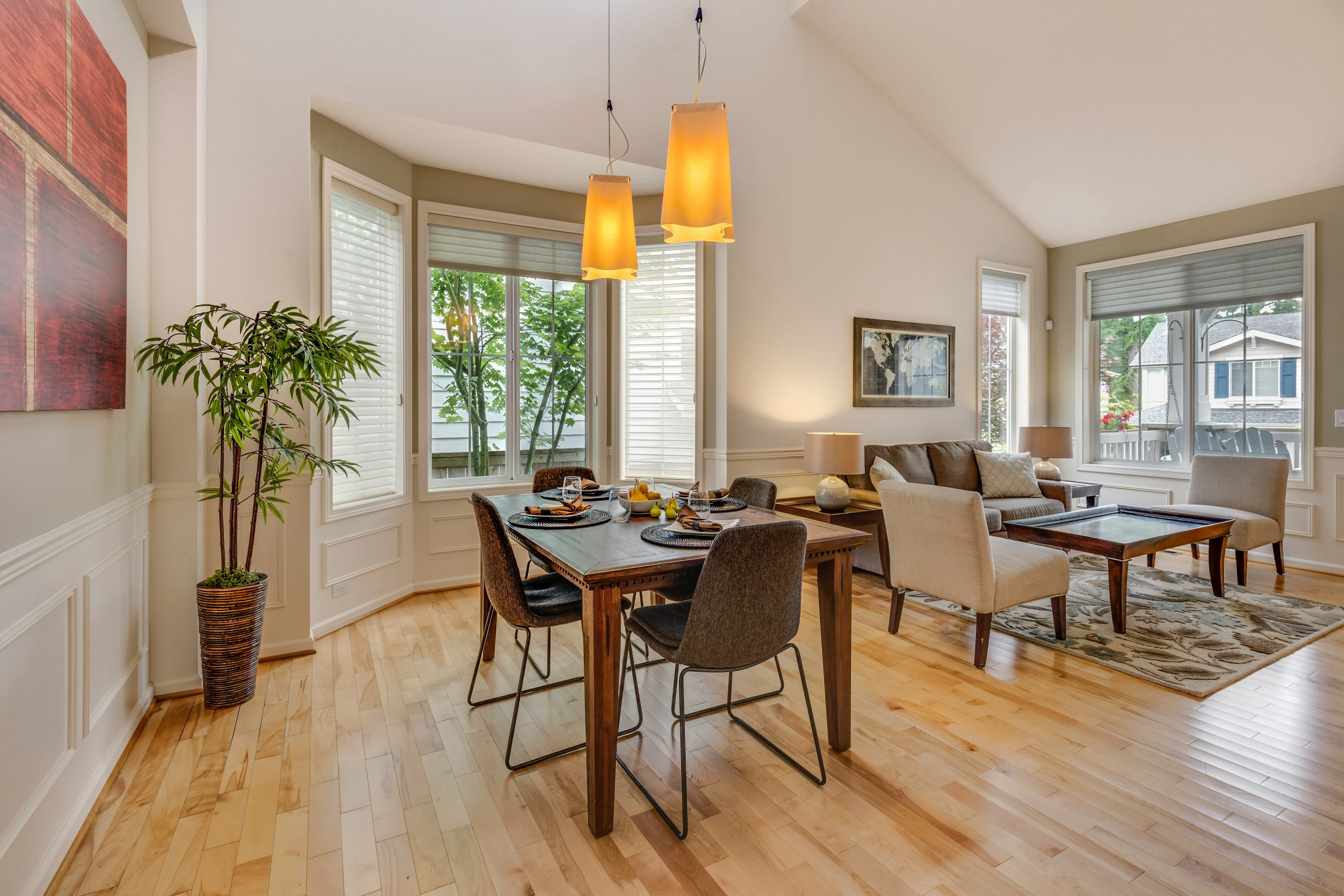 Open-plan dining and living room with wooden dining table, brown upholstered chairs, cream sofa, two orange pendant lights, potted plant, hardwood flooring, and large windows with shutters overlooking trees